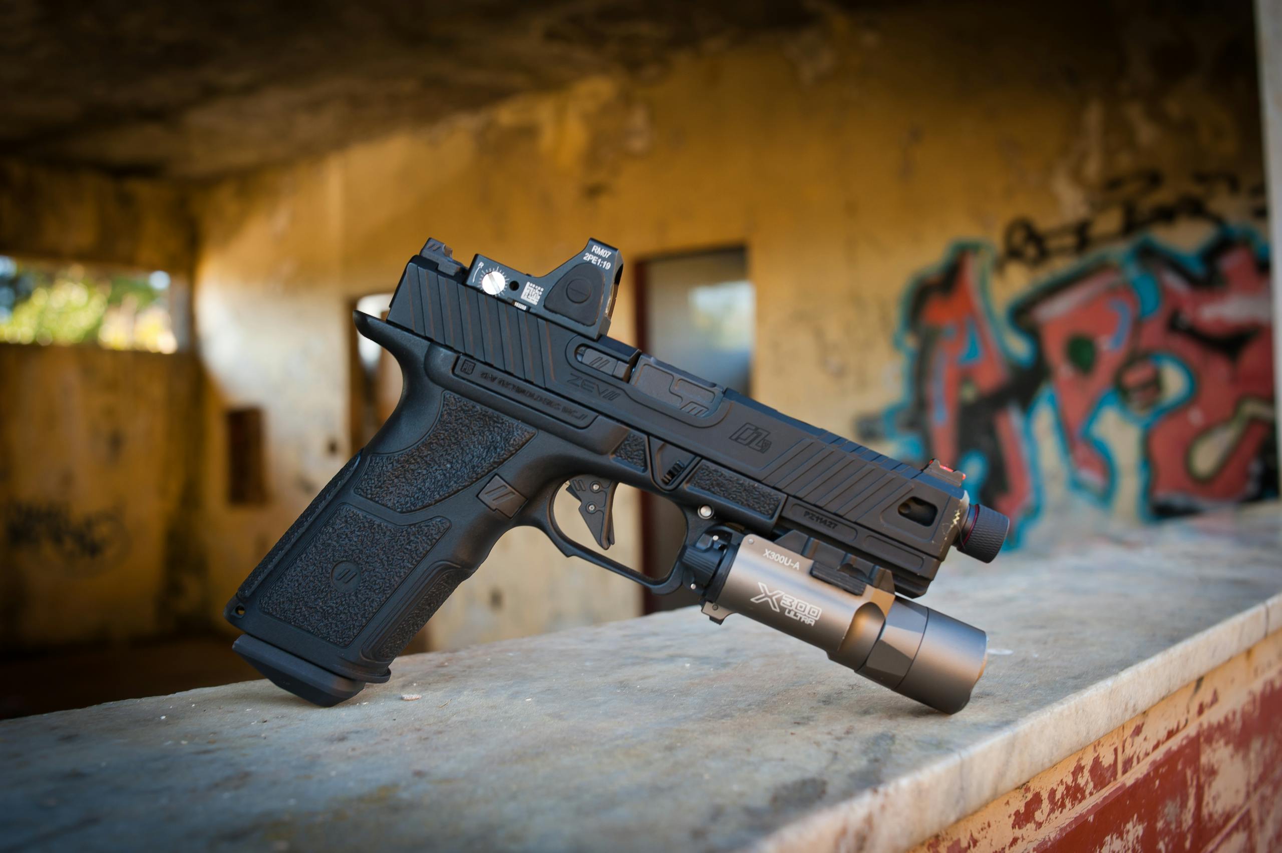 A modern black handgun with flashlight attachment on concrete in graffiti-marked urban setting.