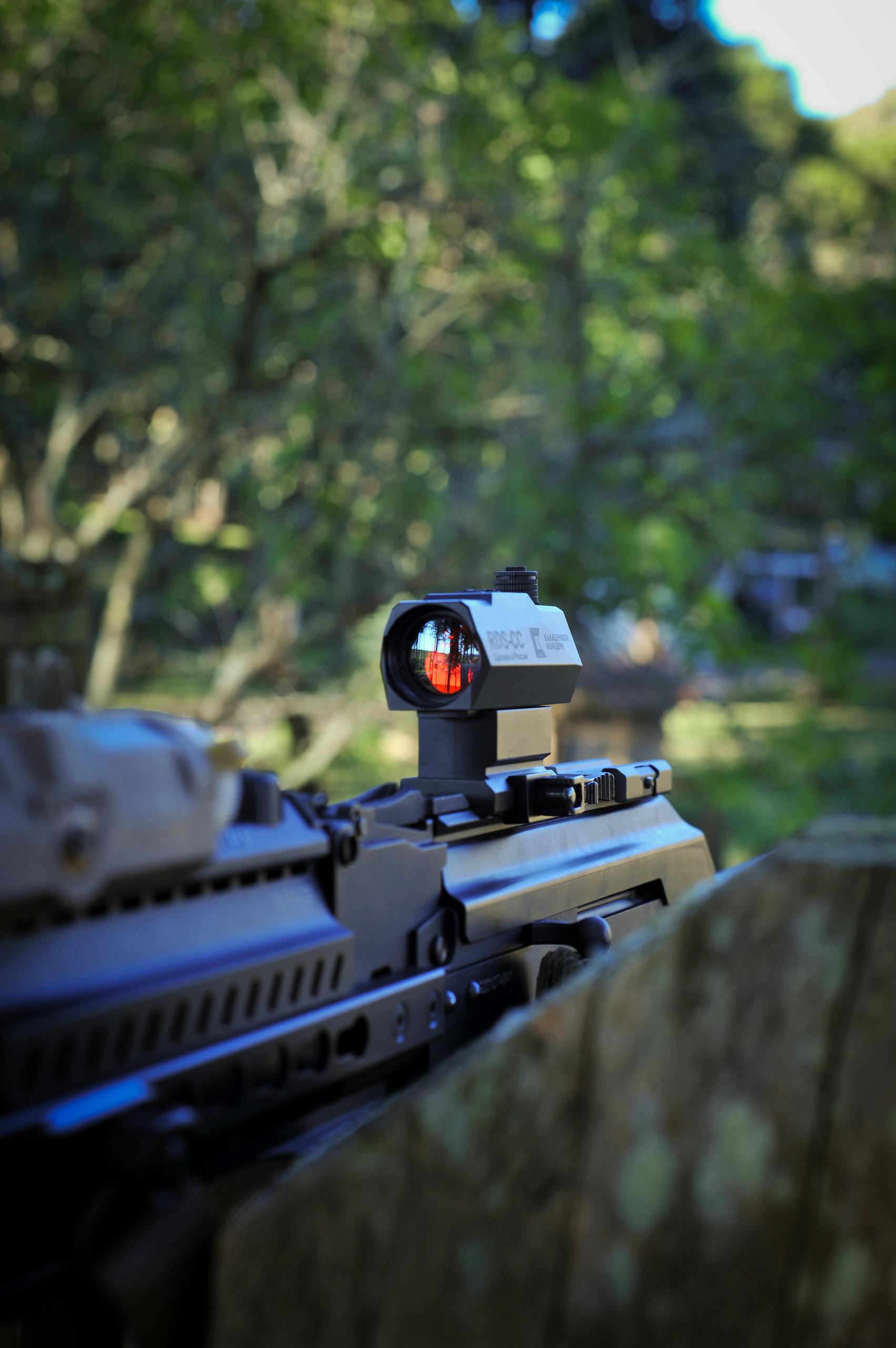 Close-up of a tactical rifle equipped with a red dot sight in an outdoor forest environment.